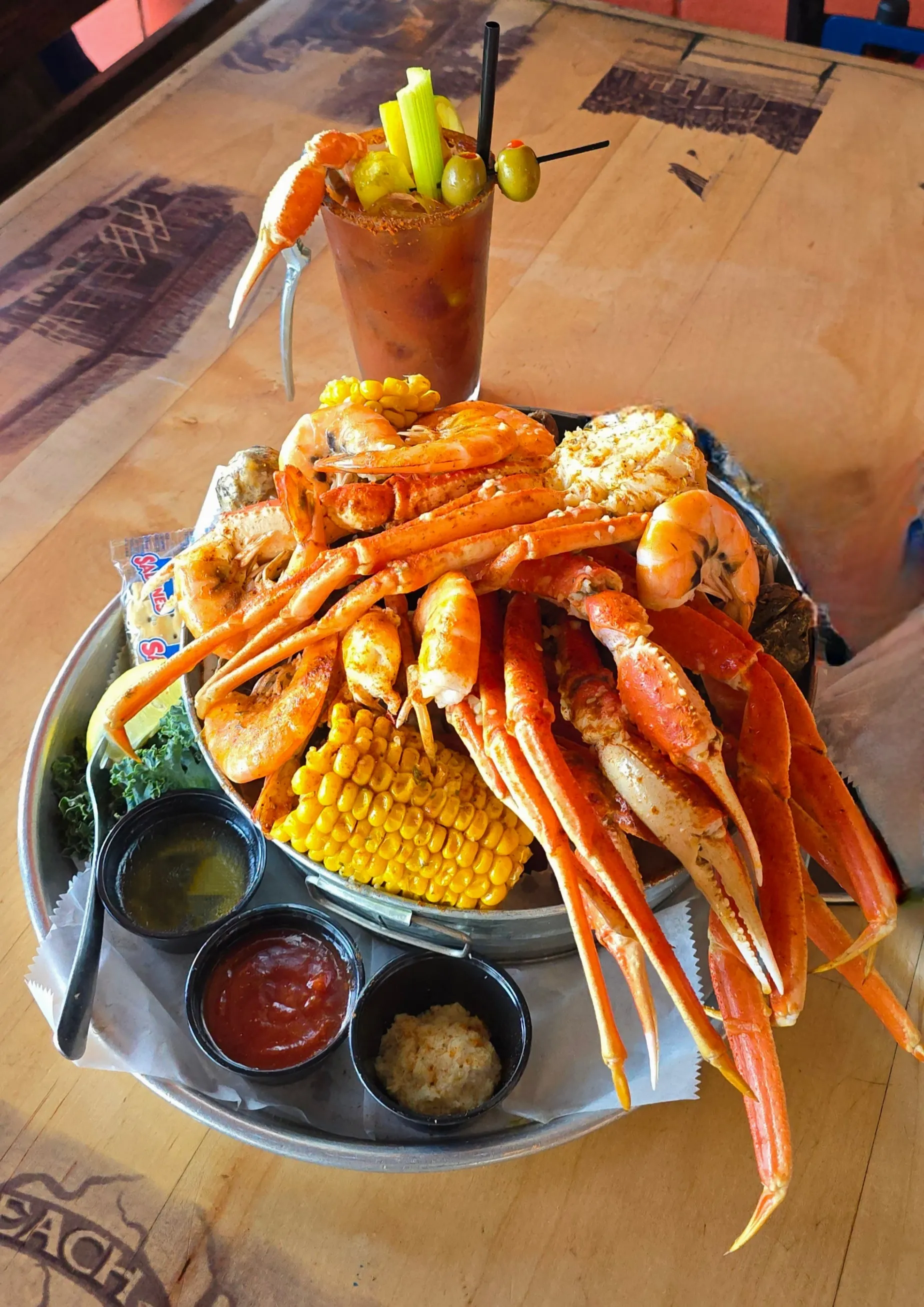 Overflowing Crab Shacks seafood bucket — snow crab legs, steamed shrimp, oysters, and corn on the cob, paired with a Crabby Mary Bloody Mary garnished with a crab claw and olives.