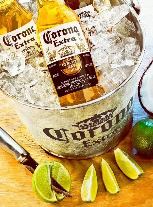 Three icy Corona Extra bottles chilling in a branded metal bucket with fresh lime wedges.